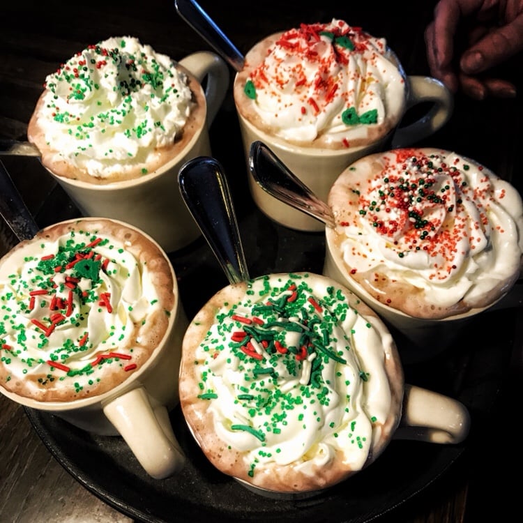 Five mugs of hot cocoa with whipped cream and sprinkles.