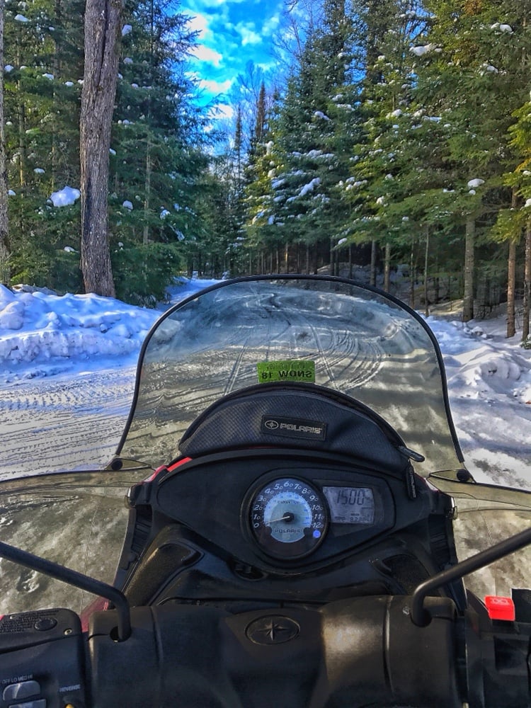 A POV shot from behind the handles of a snow mobile on a snowy trail between pine trees.