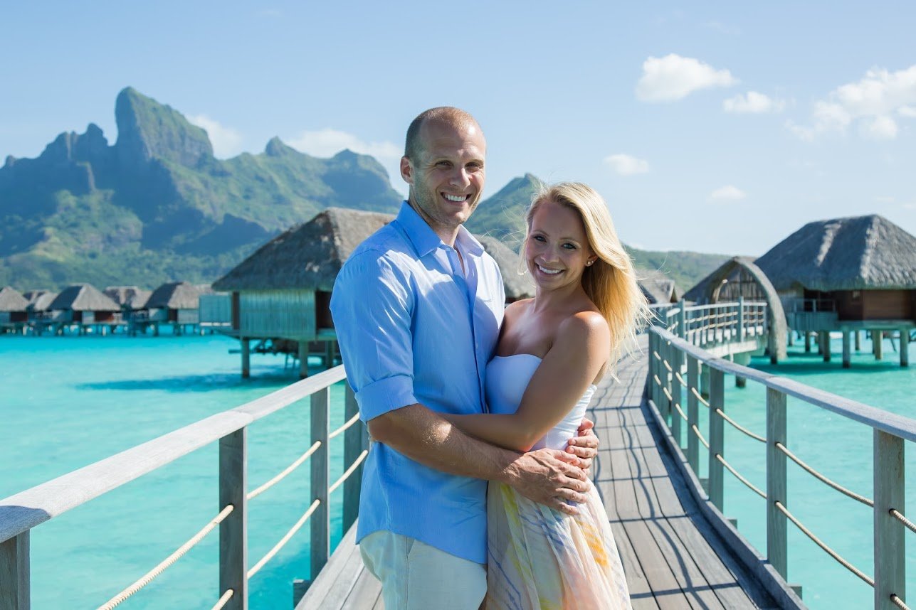Allie and her husband Kyle standing on an elevated path over tropical water leading to straw huts on stilts.