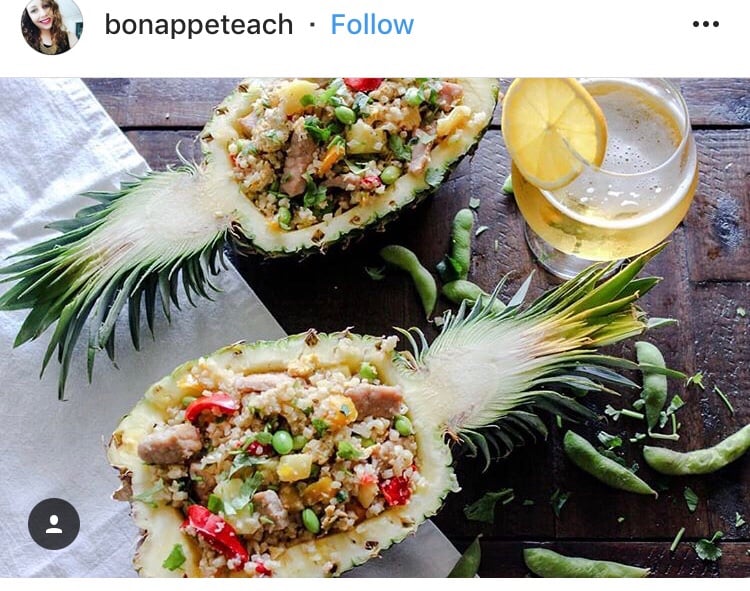 In instagram post from bonappeteach. It features a pineapple sliced in half and each half is filled with cauliflower fried rice. There is a drink with a lemon wedge next to the pineapples.