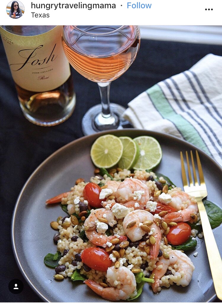 An Instagram post from hungrytravelingmama. It features shrimp and couscous on a plate next to a glass of wine.