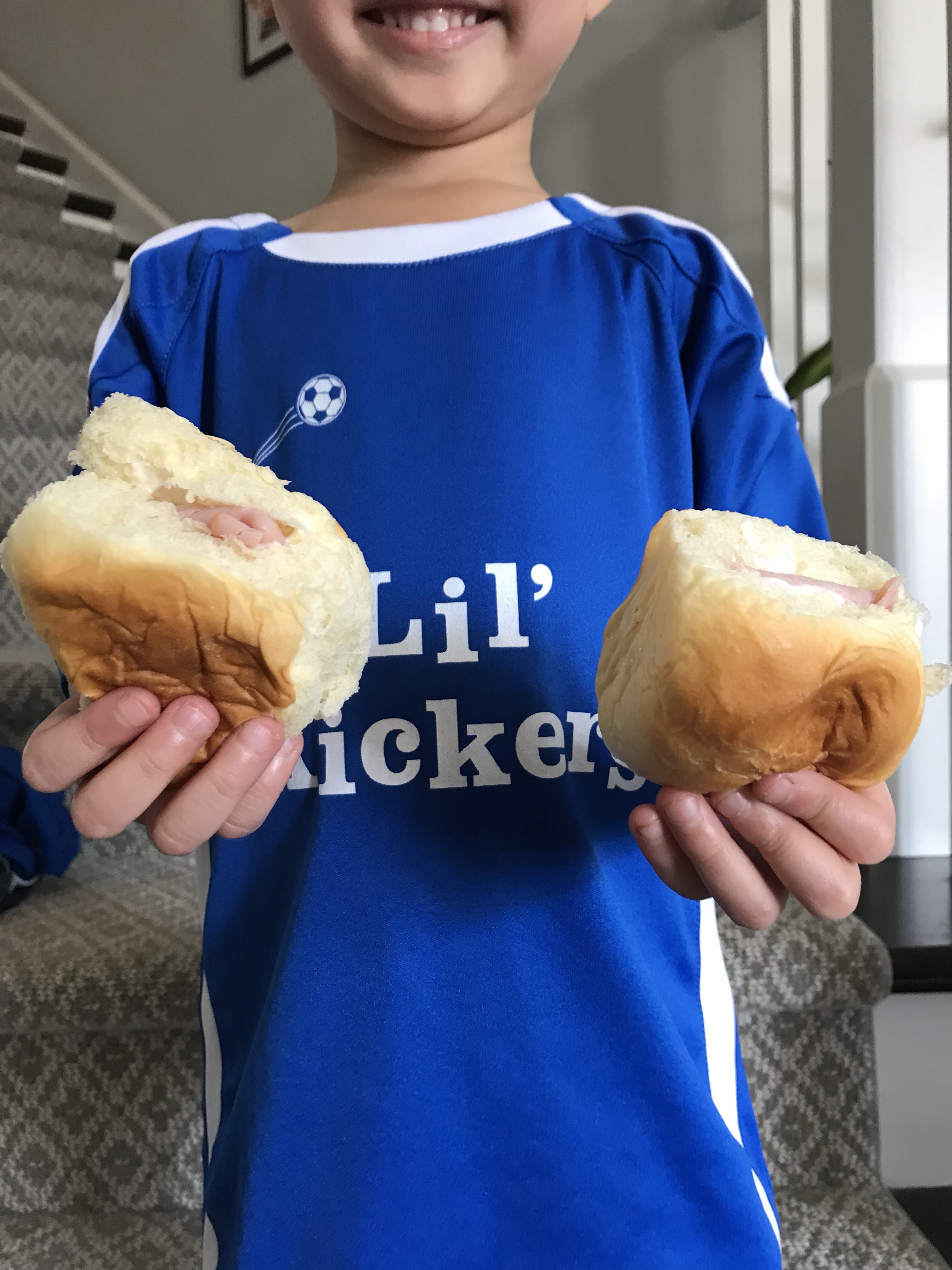 Two ham slider sandwiches being held in each hand by a boy wearing a Lil' Kickers jersey.