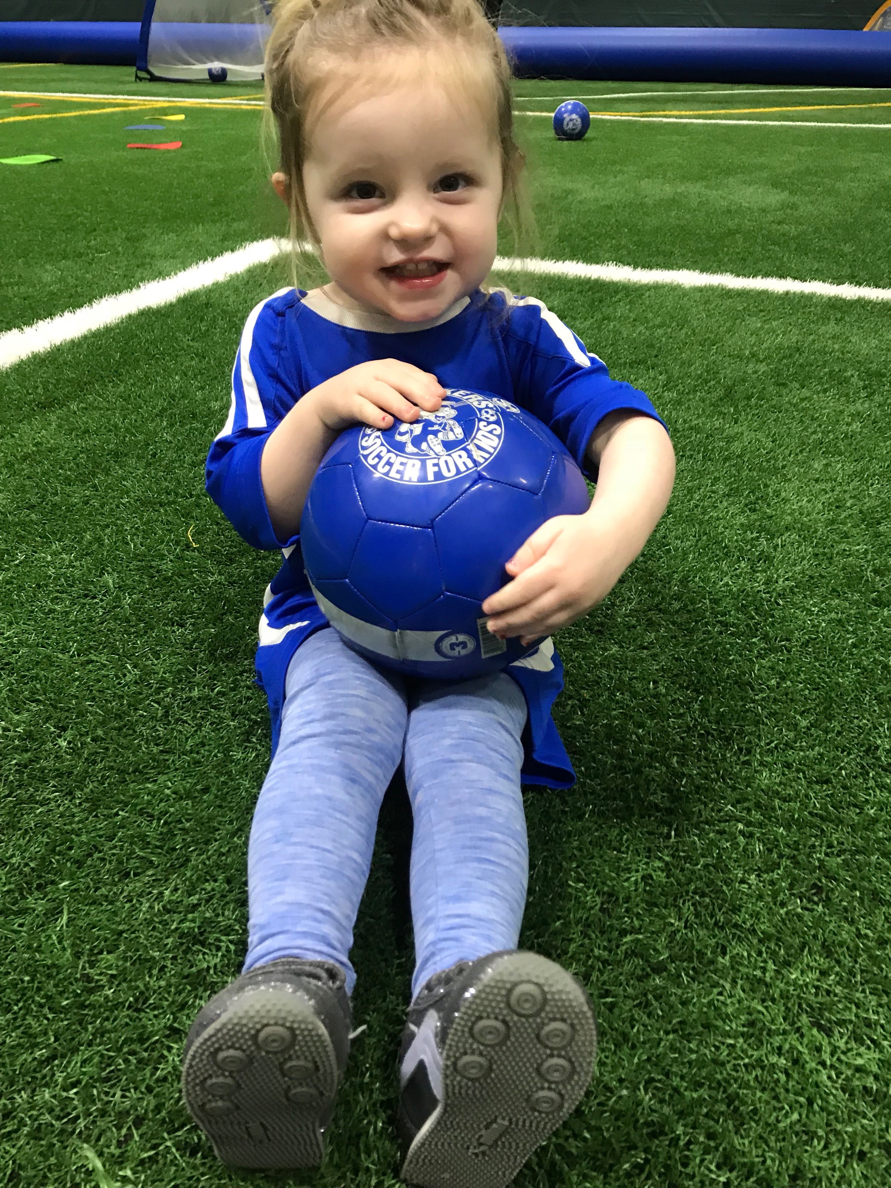 A little girl in a soccer uniform sitting on a field, holding a blue soccer ball.