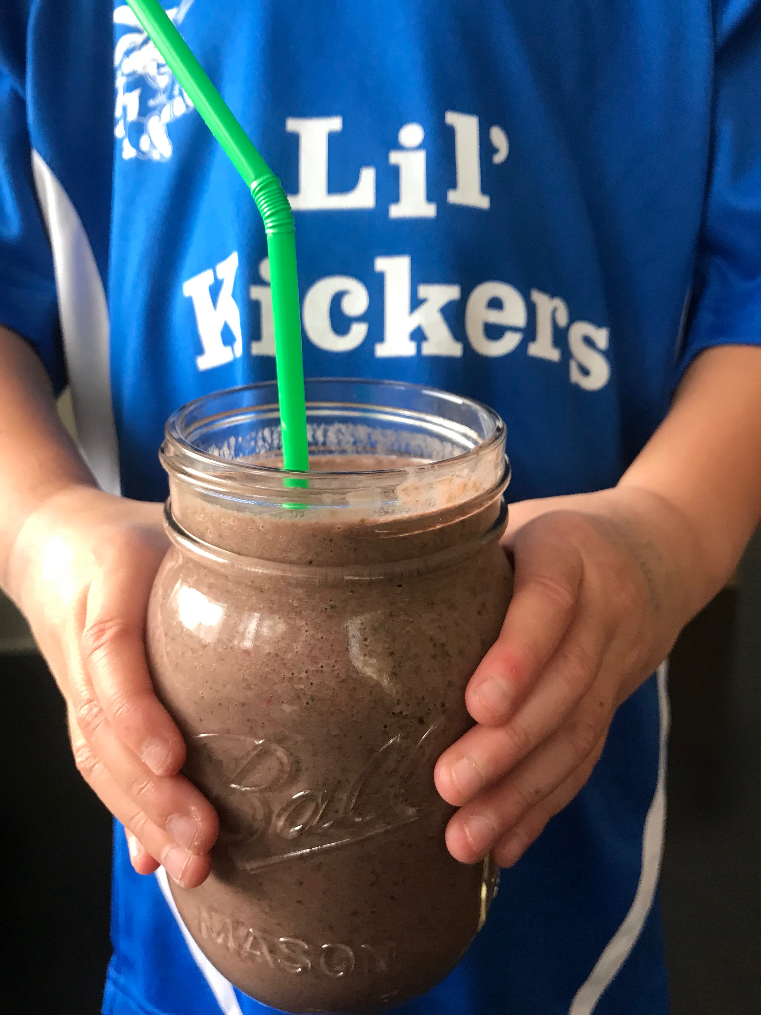 A purpleish smoothie in a mason jar with a green straw being held be a child wearing a "Lil' Kickers" shirt.