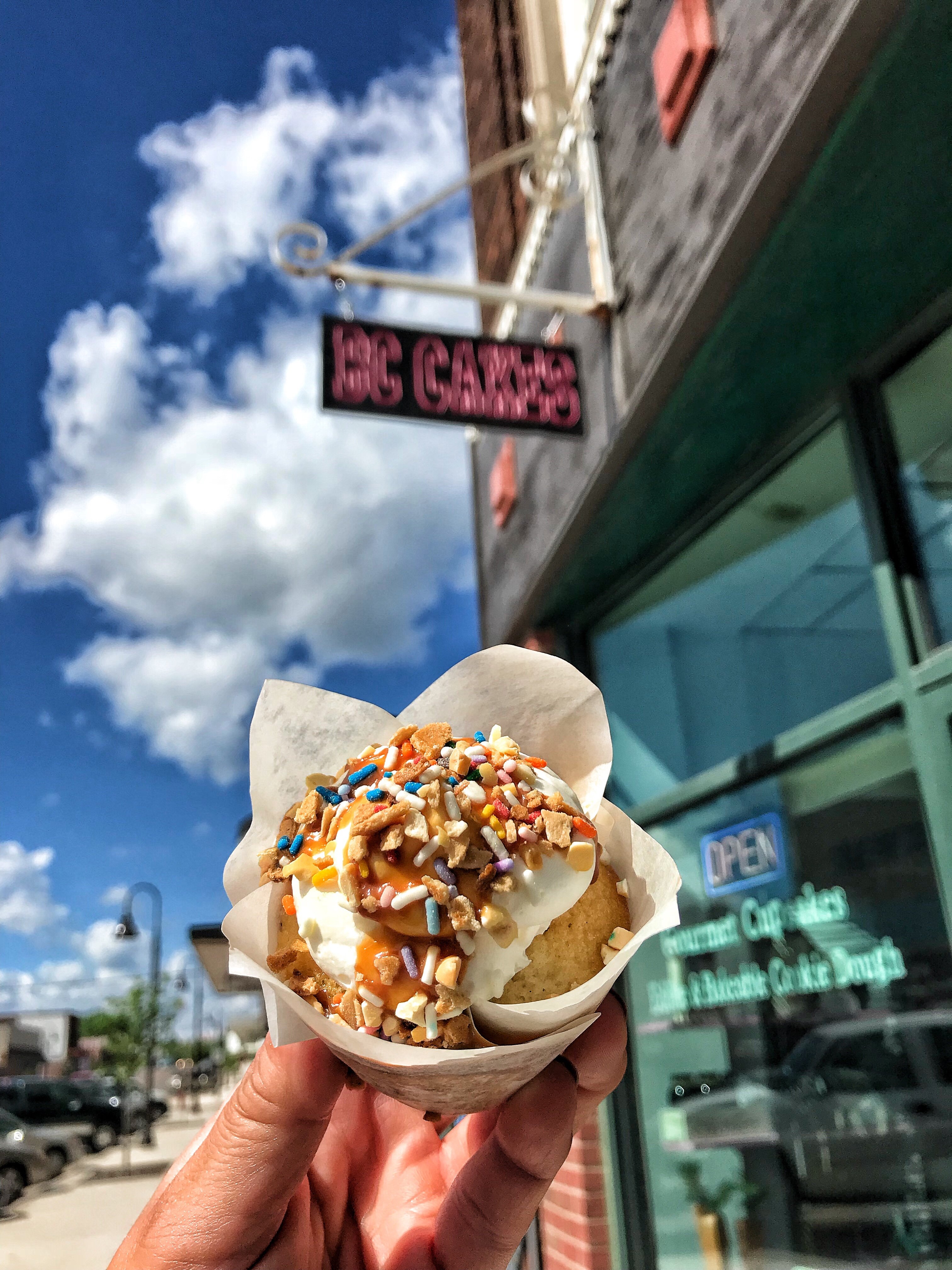 A cupcake with frosting and sprinkles being held outside under a beautiful blue and white sky.