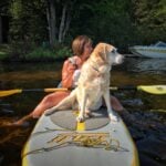 A golden retriever sitting on a paddle board in front of a woman holding a drink.