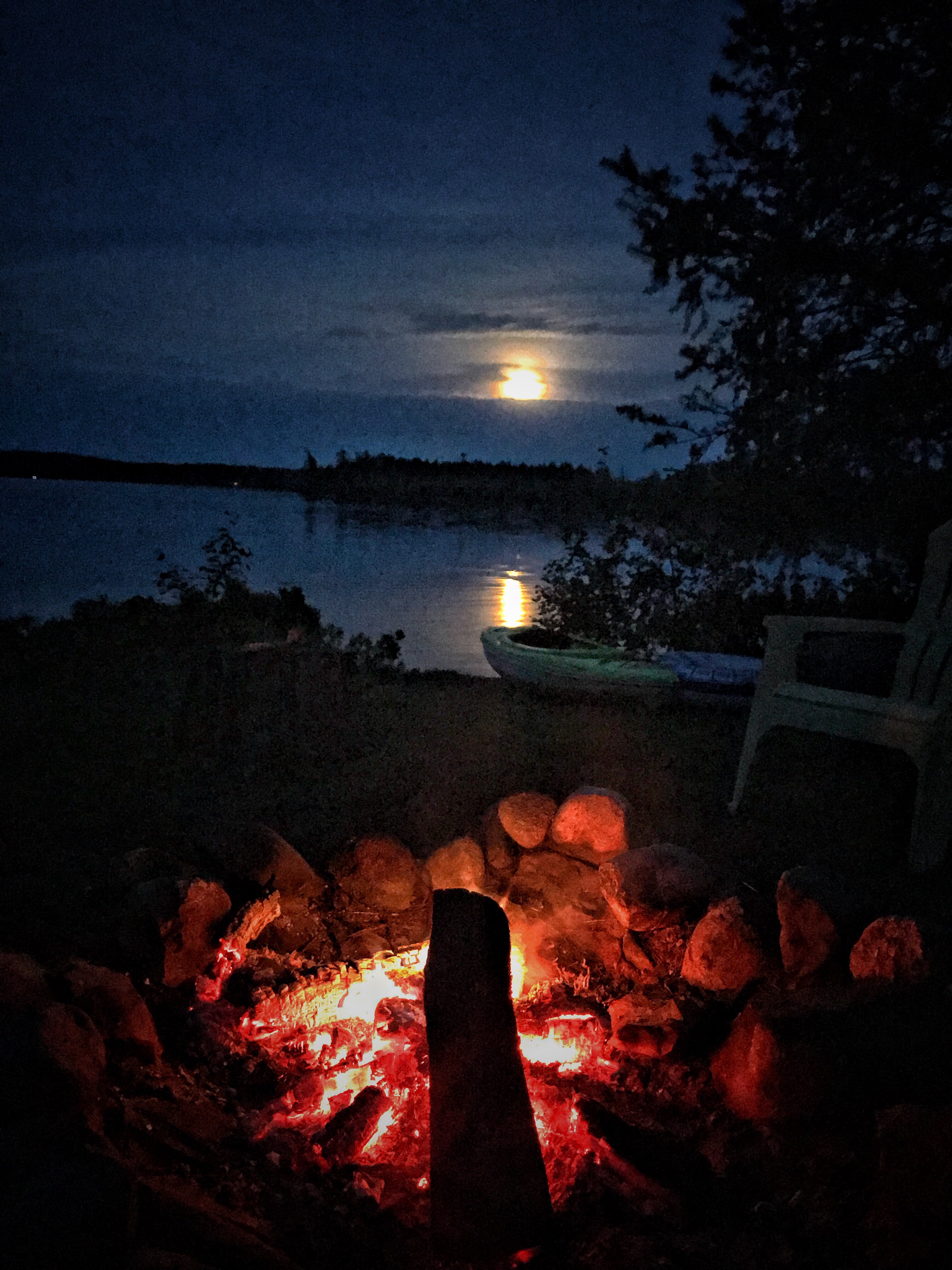 A campfire glowing at night next to a lake. A full moon shines in the sky, reflected in the water.
