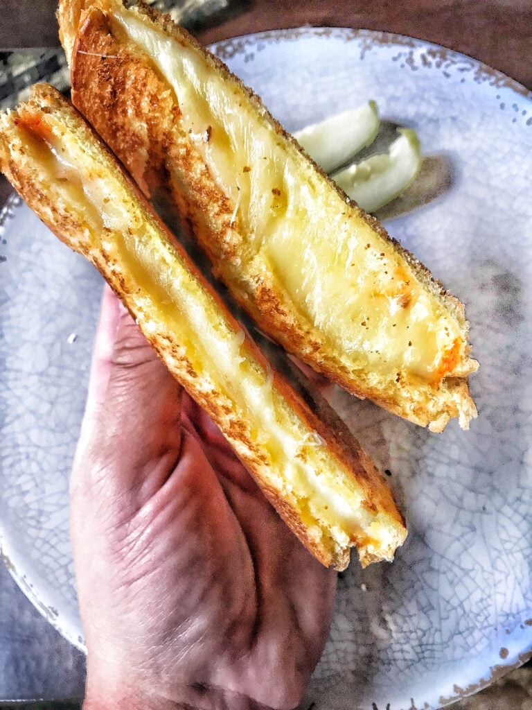 A grilled cheese sliced in half with both halves being held over a plate with pickles on it.