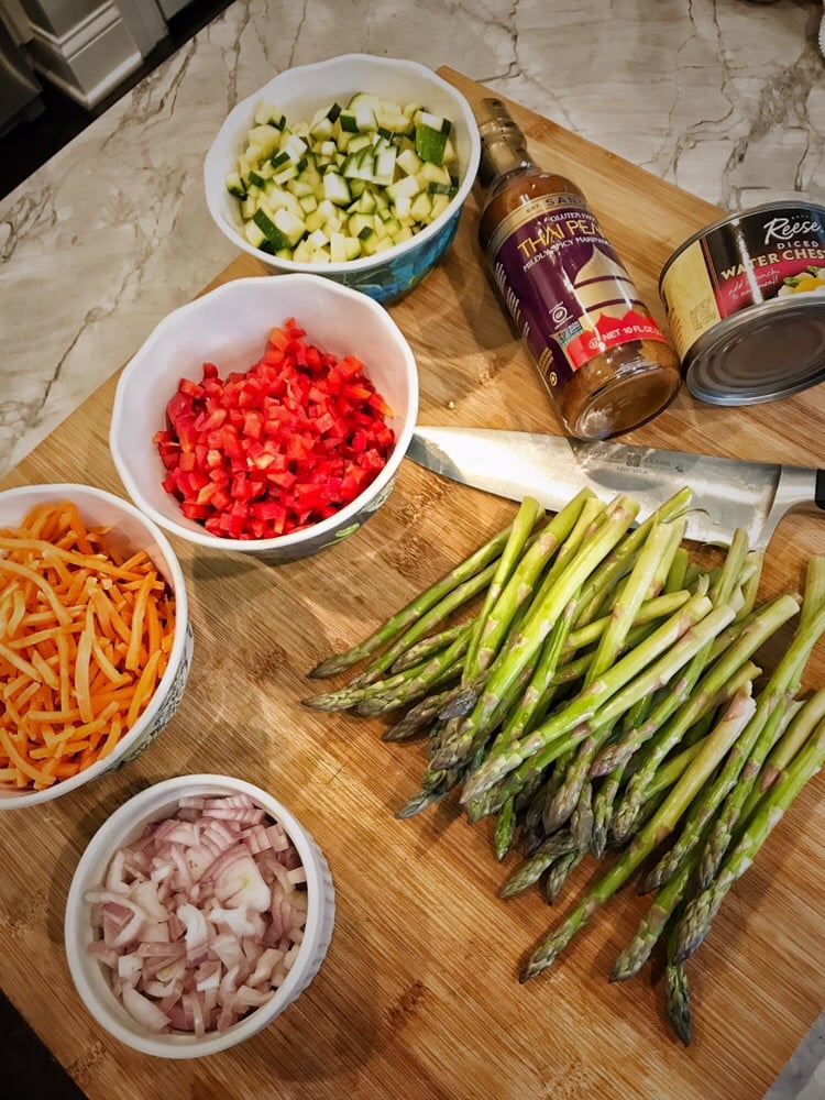 A cutting board with the ingredients for Thai lettuce wraps, including asparagus, chopped onion, carrot, zucchini, and diced red pepper next to a can of water chestnuts and Thai peanut sauce.