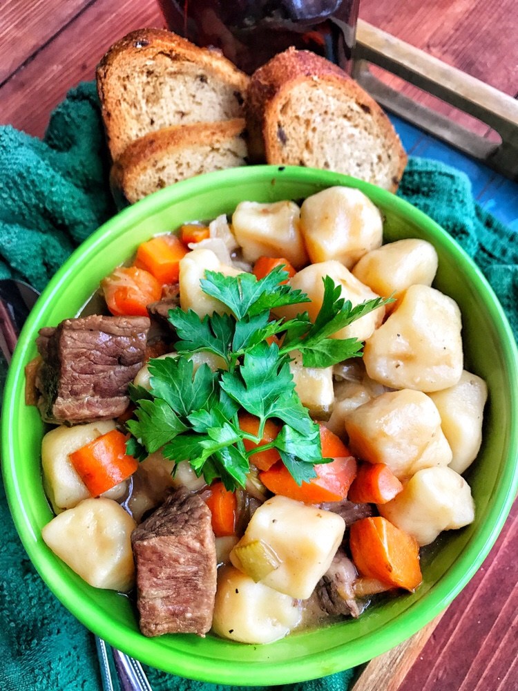 Beef cubes and carrots with mini potato dumplings in a bowl next to sliced bread.