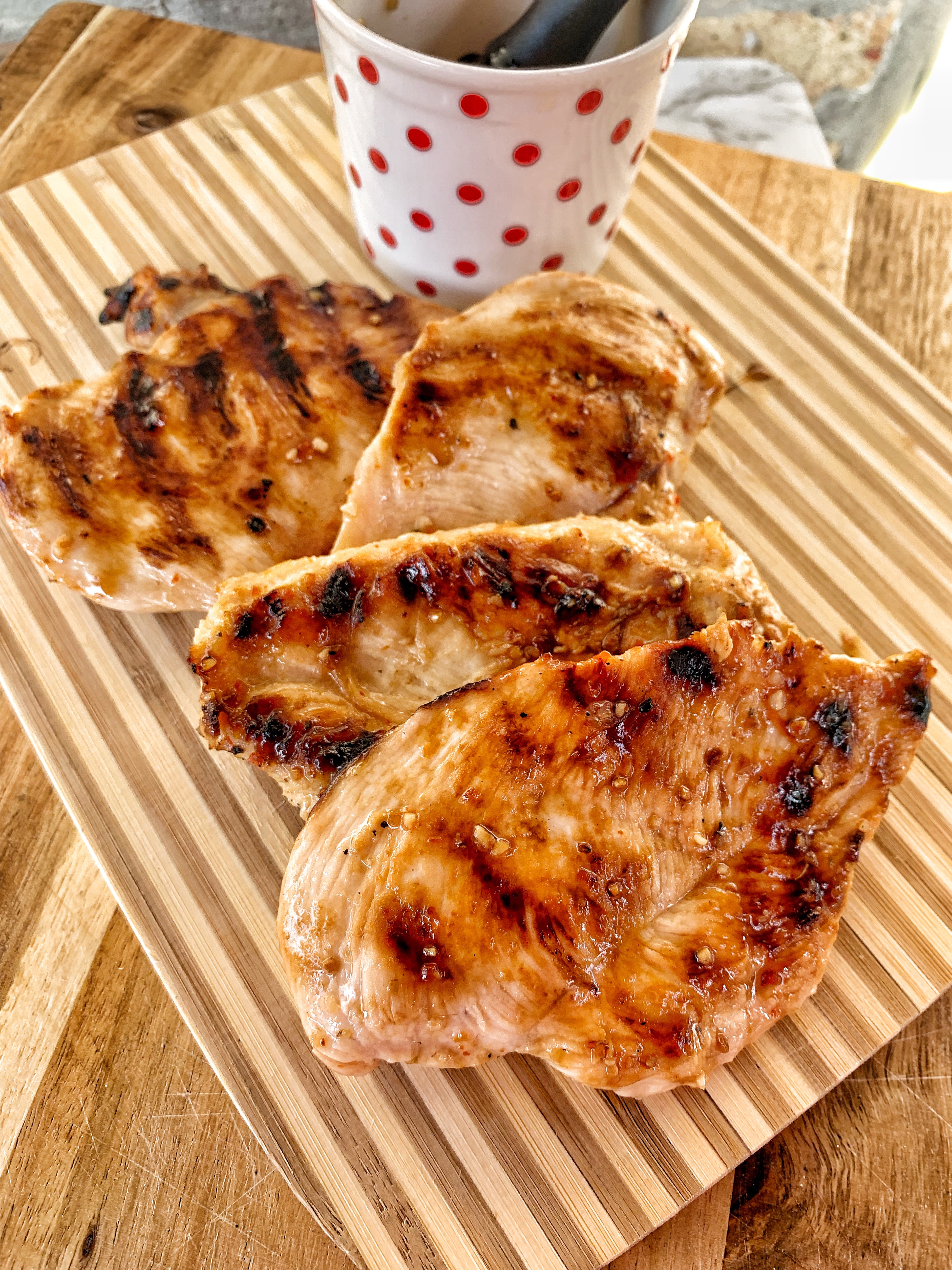 3 cooked chicken breasts with grill marks and a white cup with red polak dots that has italian dressing in it.