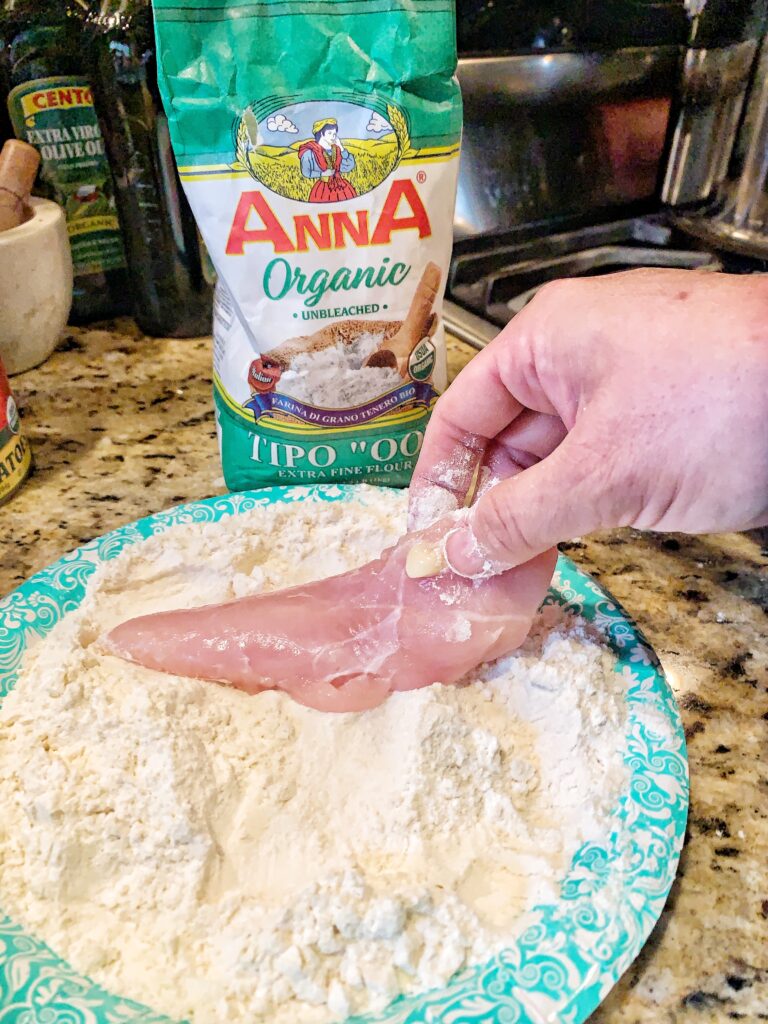 A raw chicken breast being dipped in flour on a plate