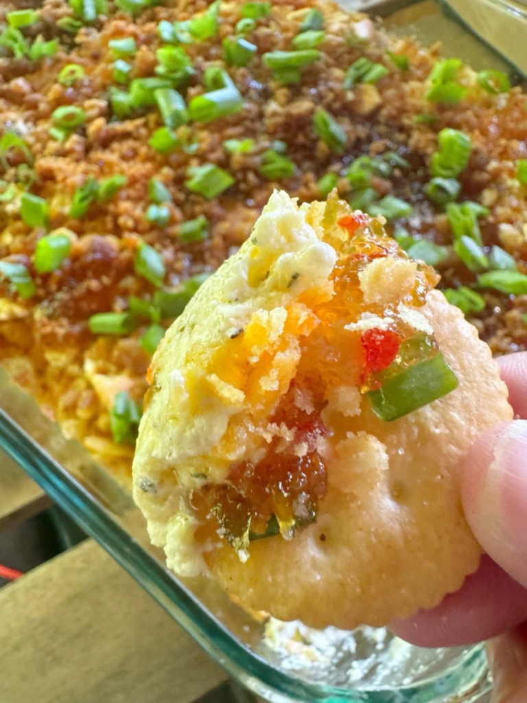 Hot cheesy party dip topped with pepper jelly, bacon, and green onions, served with crackers.