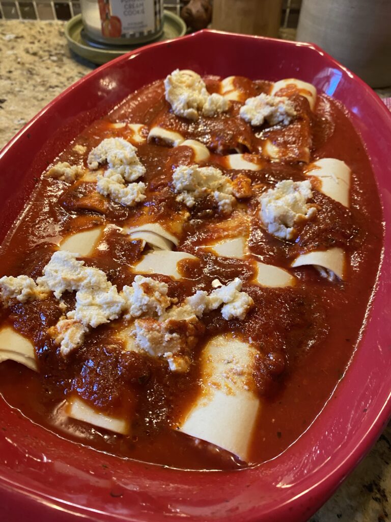 Rolled manicotti in a baking dish topped with marinara and ricotta cheese