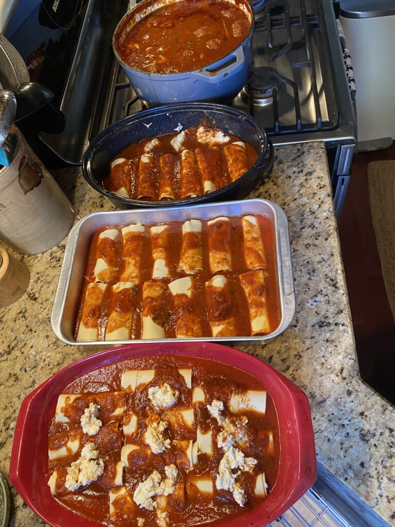 Three baking dishes of baked manicotti on the counter next to a big pot of sauce on a stove top