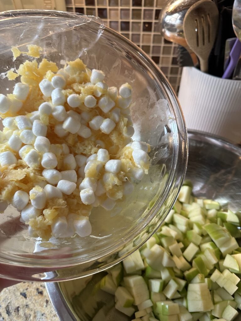 Mini marshmallows with crushed pineapple in a glass bowl next to a metal bowl of cut apples