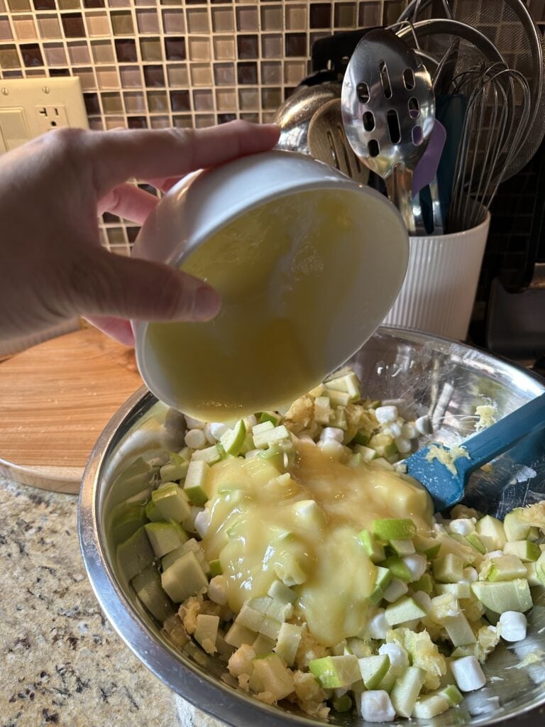 Pudding being poured over cut up apples and marshmallows in a metal bowl