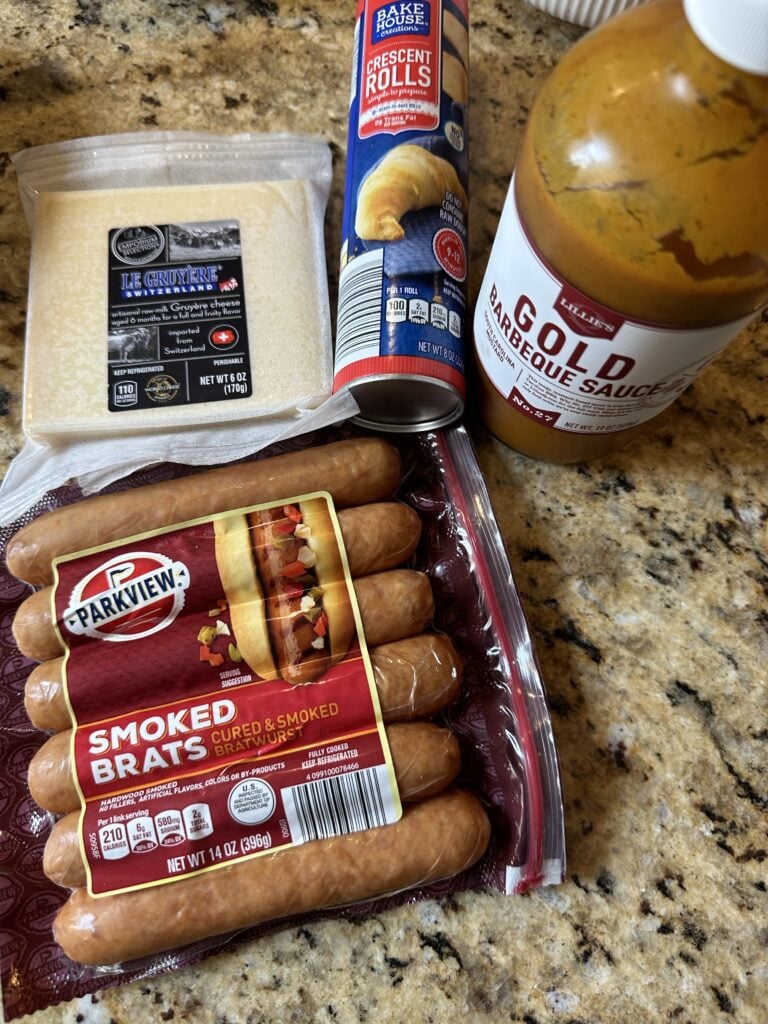 The components for the frank and Swiss bites including gruyere cheese, a crescent roll tube, gold barbeque sauce, and smoked brats
