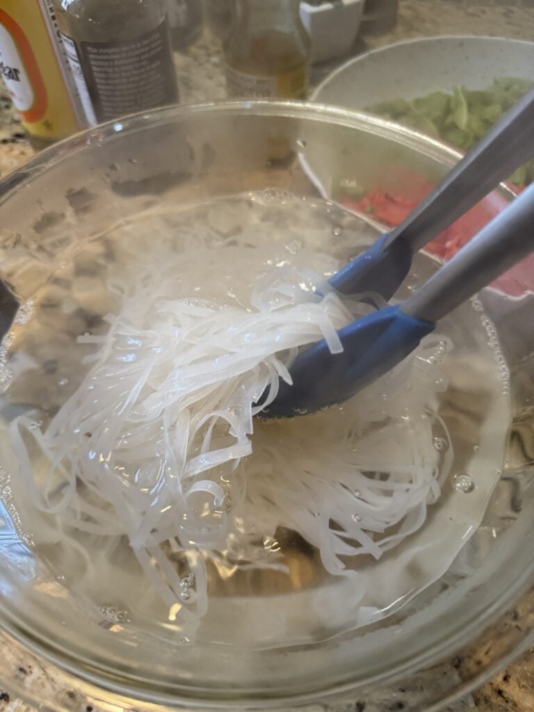 Rice noodles in a bowl of water being held up by tongs