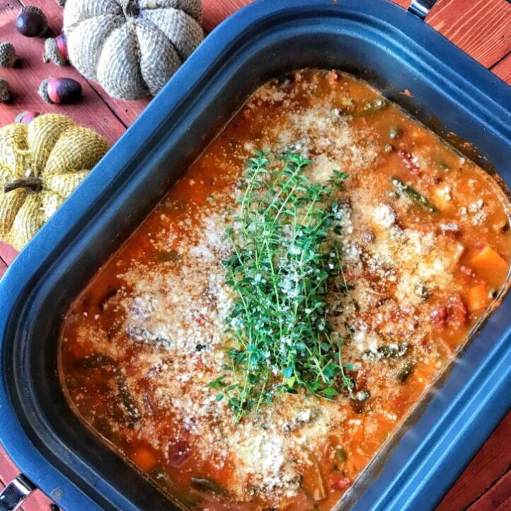 A slow cooker with a rich brownish soup broth and vegtables like carrots and beans