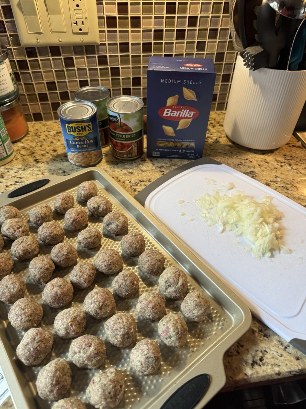 Raw meatballs on a baking tin next to grated onions on a cutting board in front of a box of Barilla medium shell pasta and a can of tomatoes and a can of cannellini beans