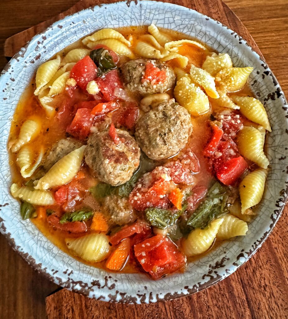 A bowl of soup showing a reddish broth, bite sized meatballs, shell pasta, tomatoes, fresh basil and a sprinkle of parmesan cheese