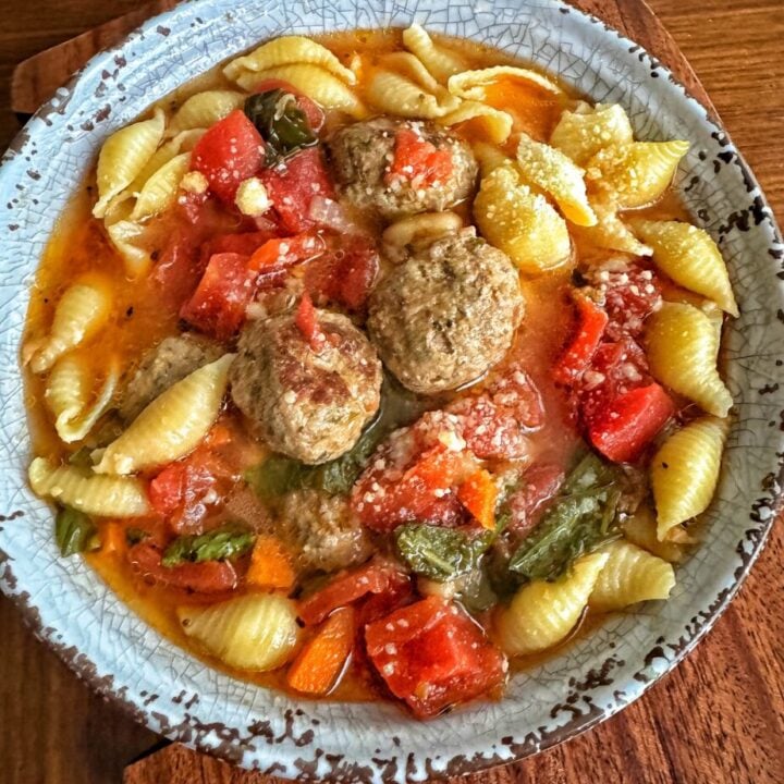 A bowl of soup showing a reddish broth, bite sized meatballs, shell pasta, tomatoes, fresh basil and a sprinkle of parmesan cheese