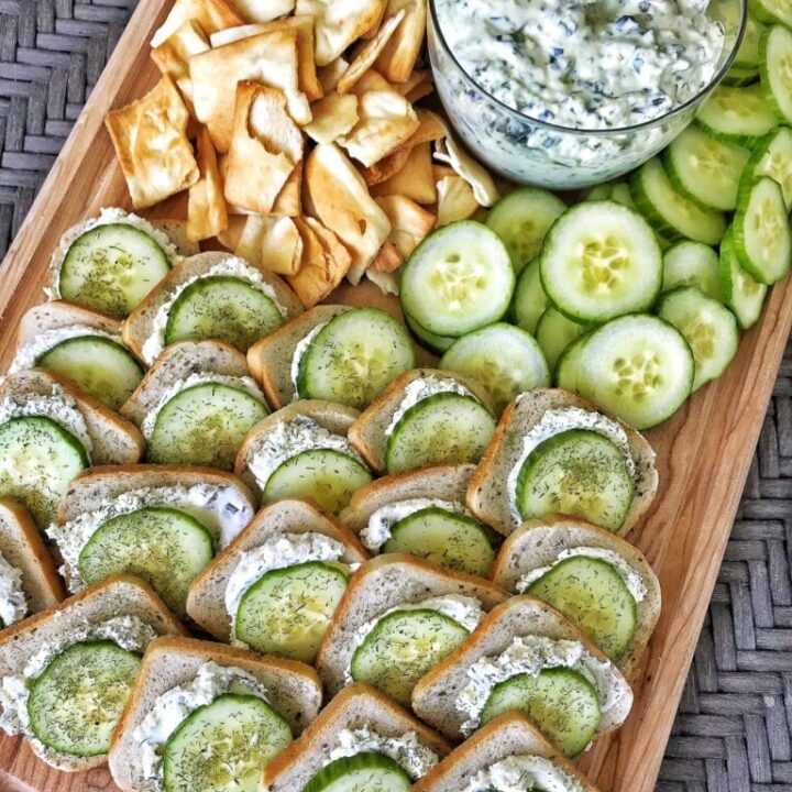 Platter of rye bread topped with cream cheese thats been seasoned as well as cucumber slices and sill. A bowl of spinch dip and pita crackers