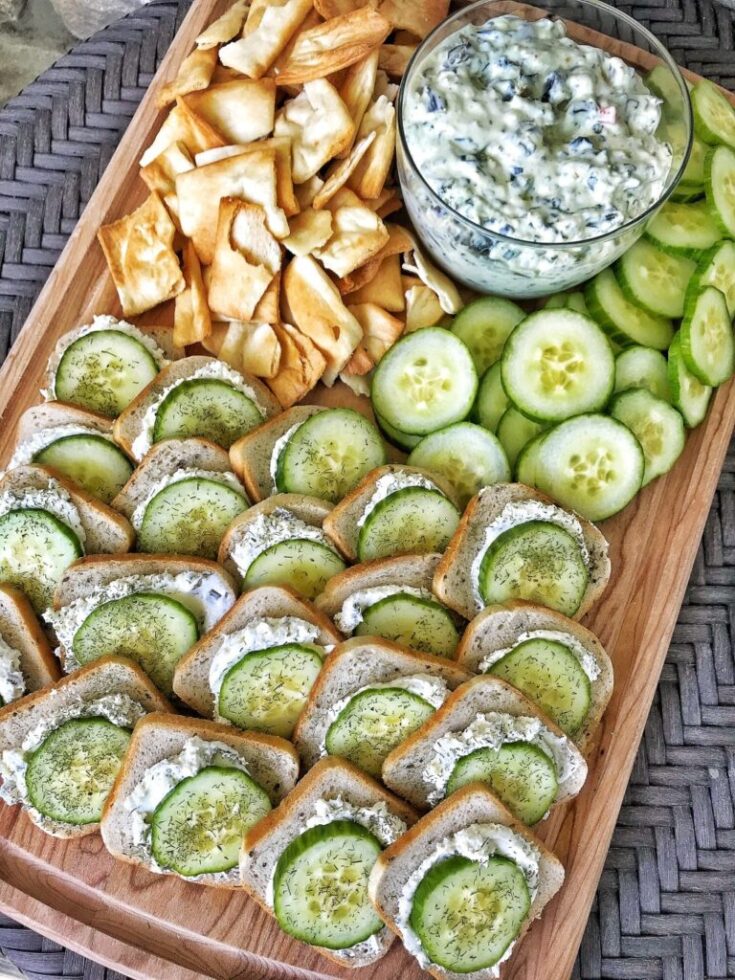 Platter of rye bread topped with cream cheese thats been seasoned as well as cucumber slices and sill. A bowl of spinch dip and pita crackers