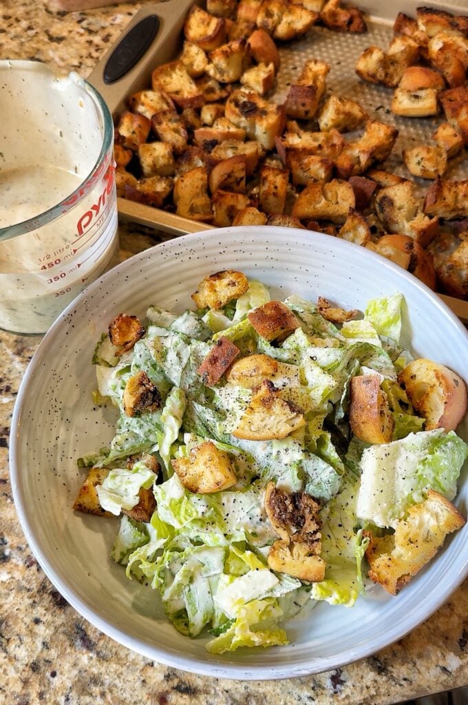 plate with romaine lettuce, homemade bread croutons, homemade dressing and a tray of croutons behind the plate