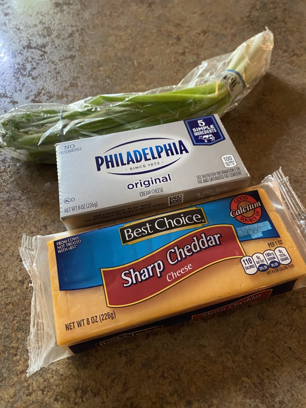 A package of green onion, Philadelphia cream cheese, and a block of sharp cheddar cheese on a kitchen counter