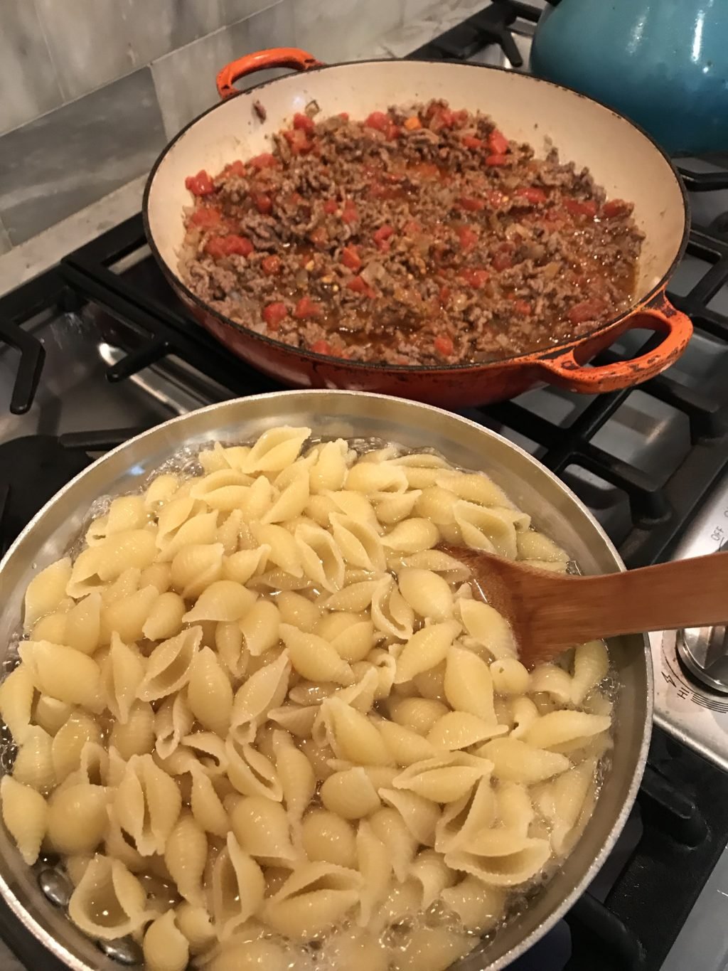 A pot of pasta water on a stove top next to a red pan of meat sauce