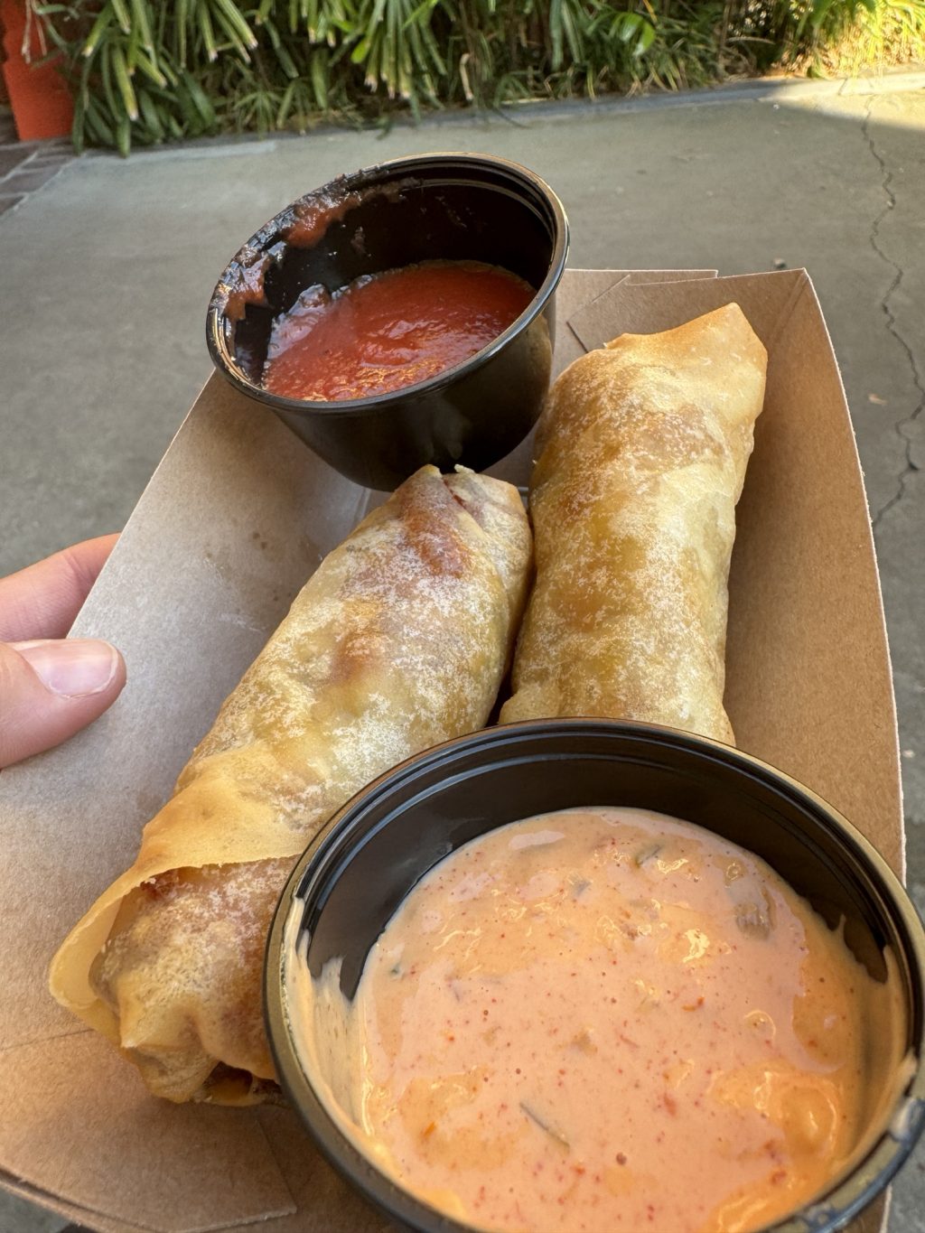 Two cheeseburger spring rolls with a red sauce and a pink sauce in an open cardboard container