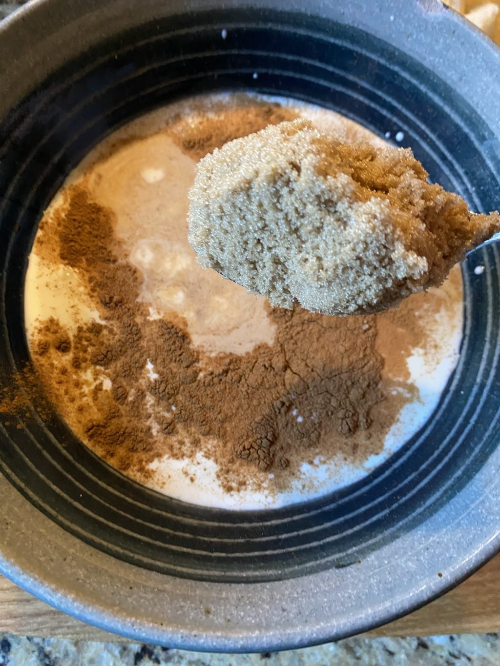 A spoon of sugar held above a bowl of French toast batter with visible cinnamon on top