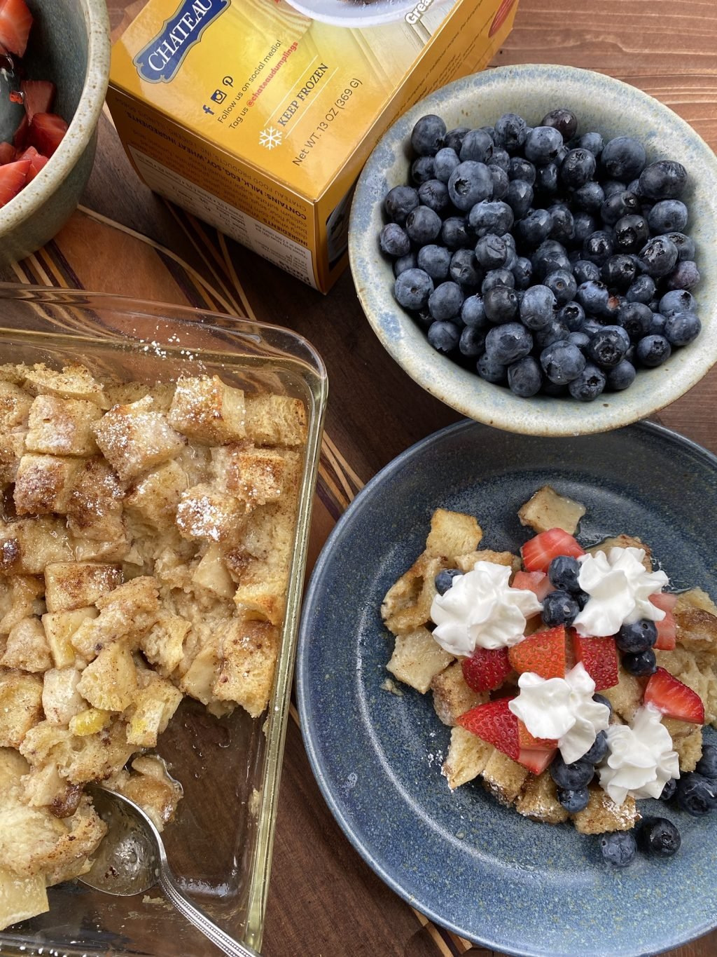 A baking dish of French toast casserole next to a plate with pieces of French toast casserole topped with berries and whipped cream alongside a bowl of blueberries and a box of Chateau Bread Dumplings