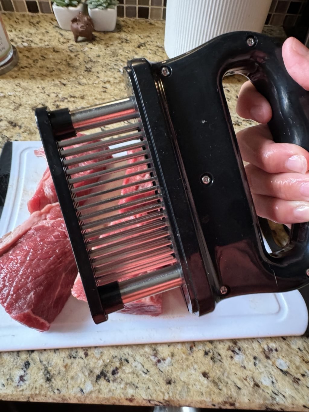 A meat tenderizer being held above chunks of beef on a white cutting board