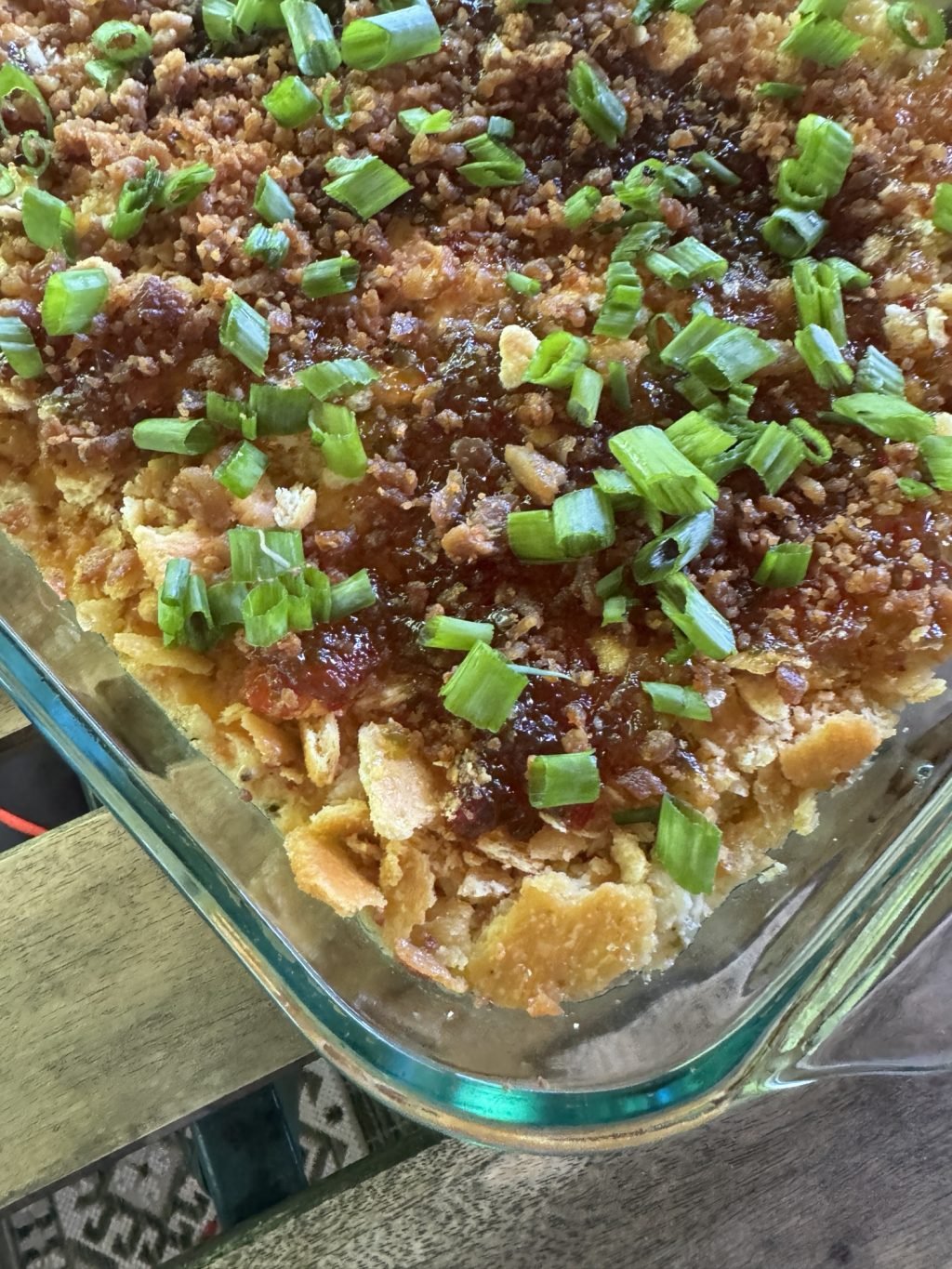 A glass baking dish of party dip with crumbled crackers, baked cream cheese, pepper jelly, and green onions.