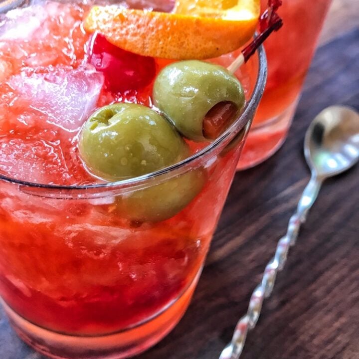 A brandy old fashioned garnished with olives, cherry, and an orange twist.