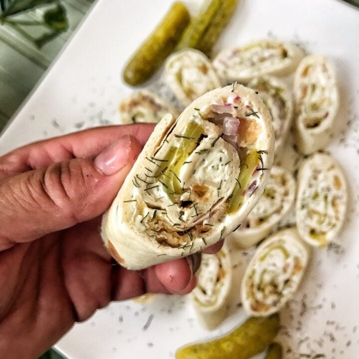 Chopped pickles, meats, and spread in a sliced wrap held above a plate of other pickle pinwheels.