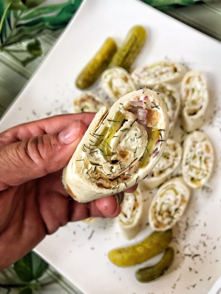Chopped pickles, meats, and spread in a sliced wrap held above a plate of other pickle pinwheels.