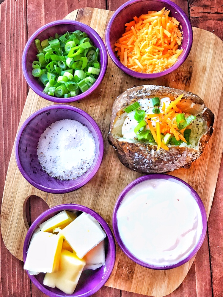 A loaded baked potato surrounded by purple cups of shredded cheese, chopped green onions, salt, sour cream, and butter pats.