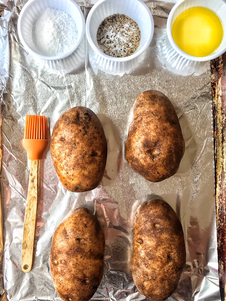 A baking pan with two potatoes, an orange kitchen brush, and three small cups of salt, pepper, and olive oil.