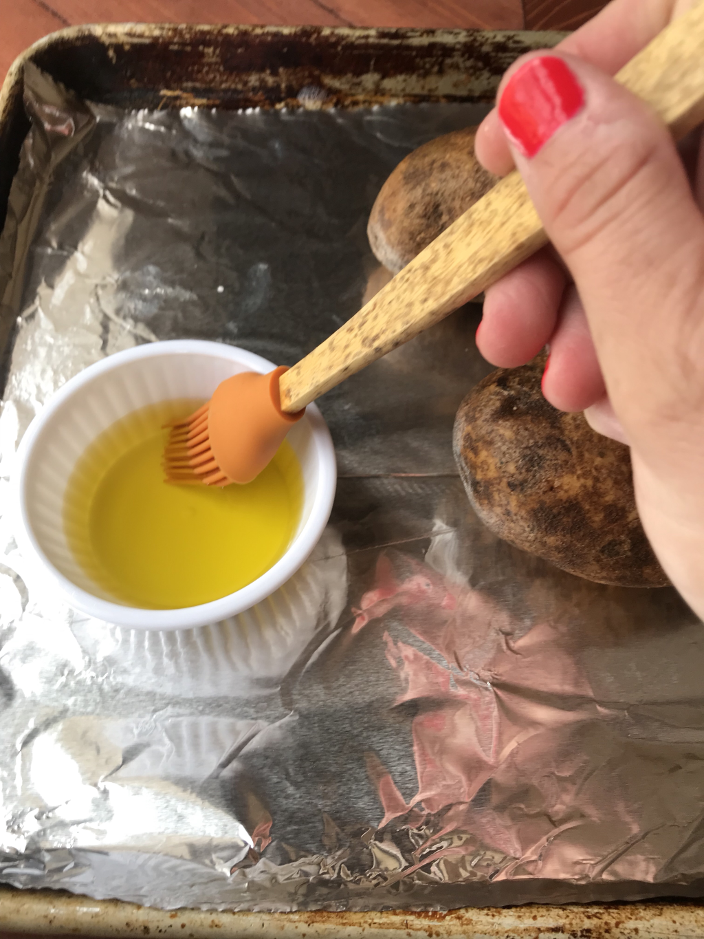 A kitchen brush being dipped into a cup of olive oil next to two potatoes on a foil baking sheet.