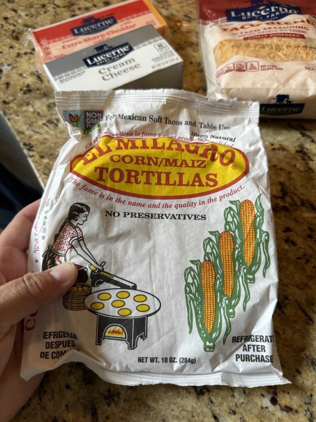 A package of corn tortillas being held above a counter that has a package of cream cheese, cheddar cheese, and shredded cheese.