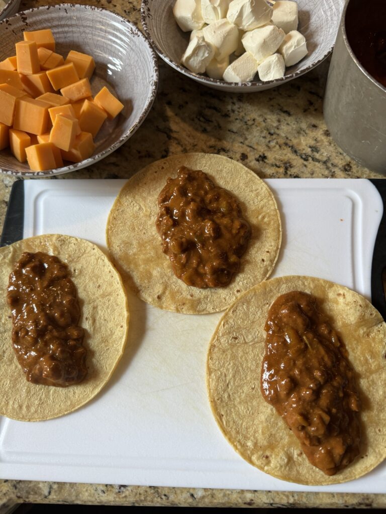 Three tortillas with chili on top on a white cutting board next to bowls of cheddar cheese and cream cheese.