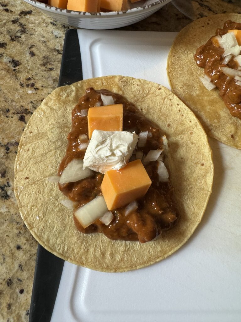 A corn tortilla with chili, diced onion, cheddar cheese cubes, and cream cheese on it.
