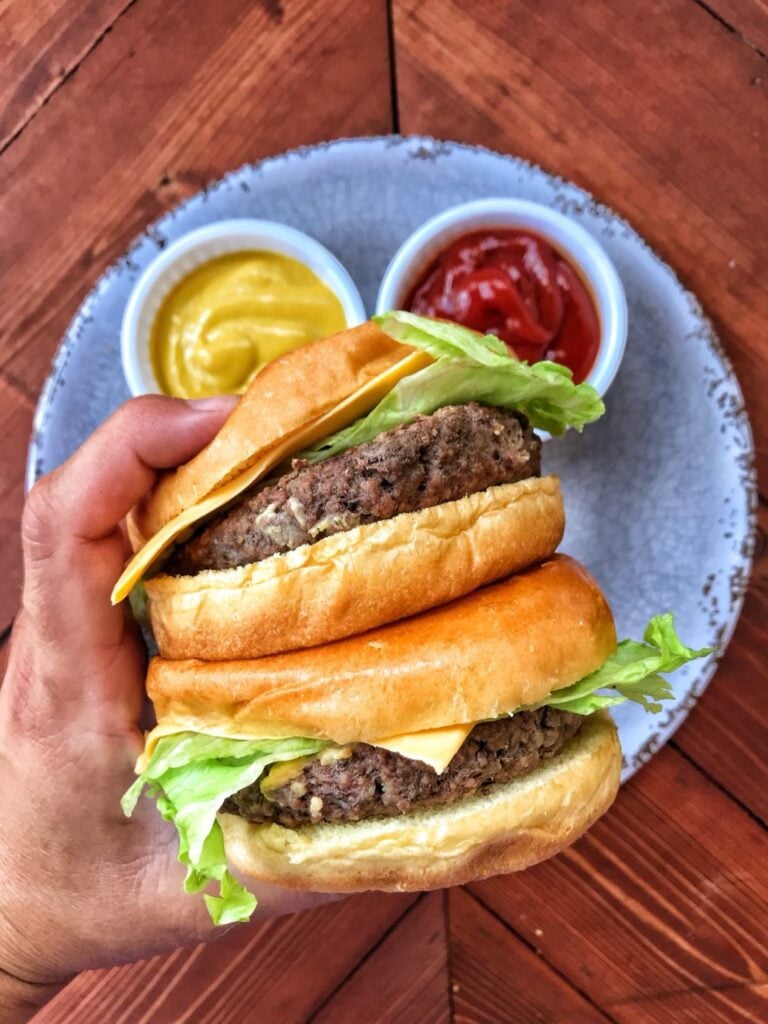 Two cheeseburgers stacked on top of each other being held above a plate with cups of ketchup and mustard.