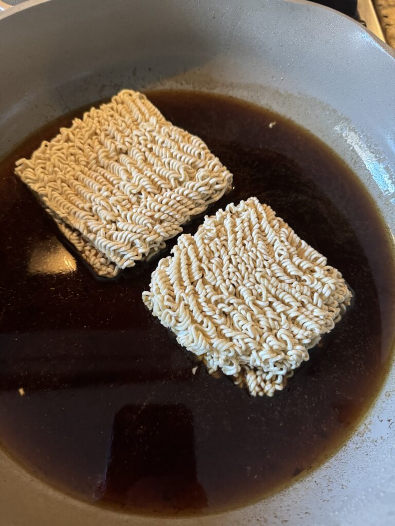 Two squares of uncooked ramen noodles in brown liquid in a pan.