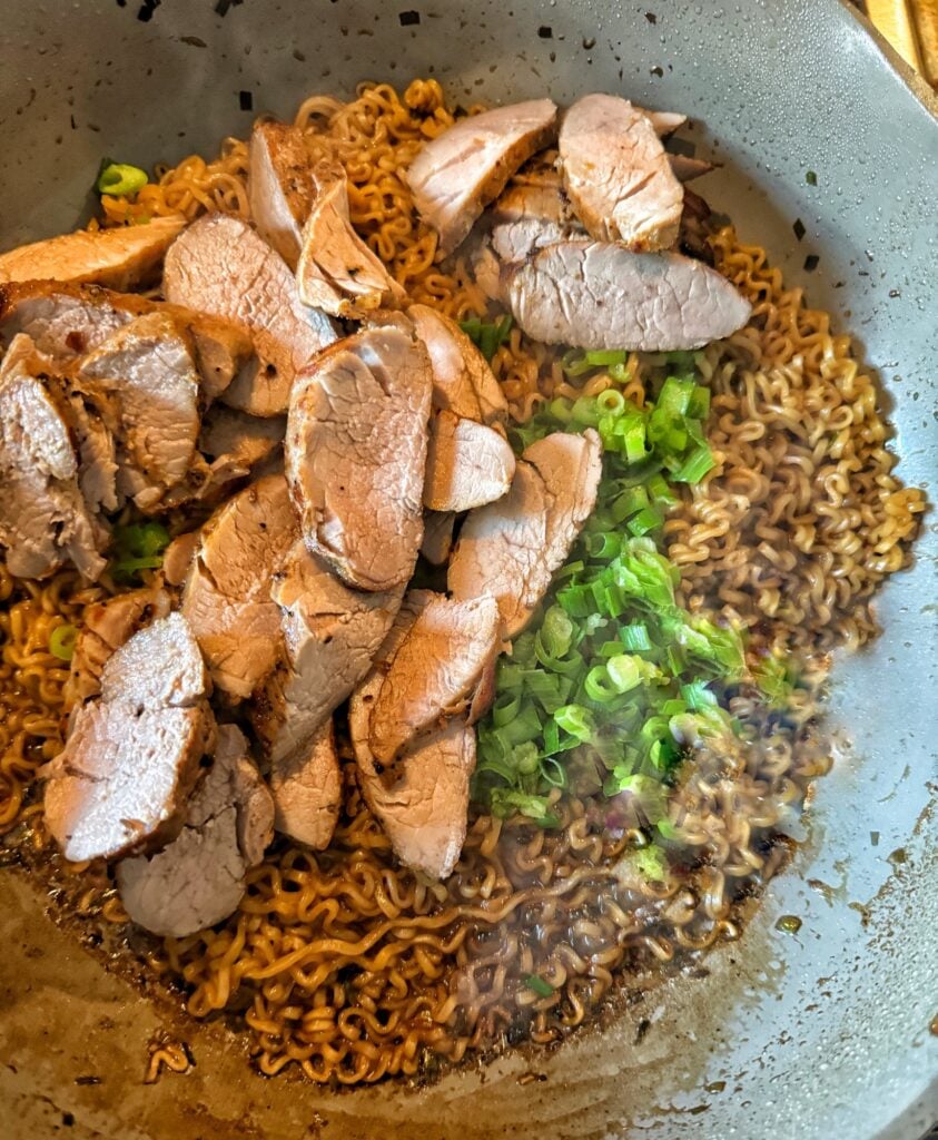 Cooked ramen noodles in brown sauce in a pan with sliced green onions and pork.
