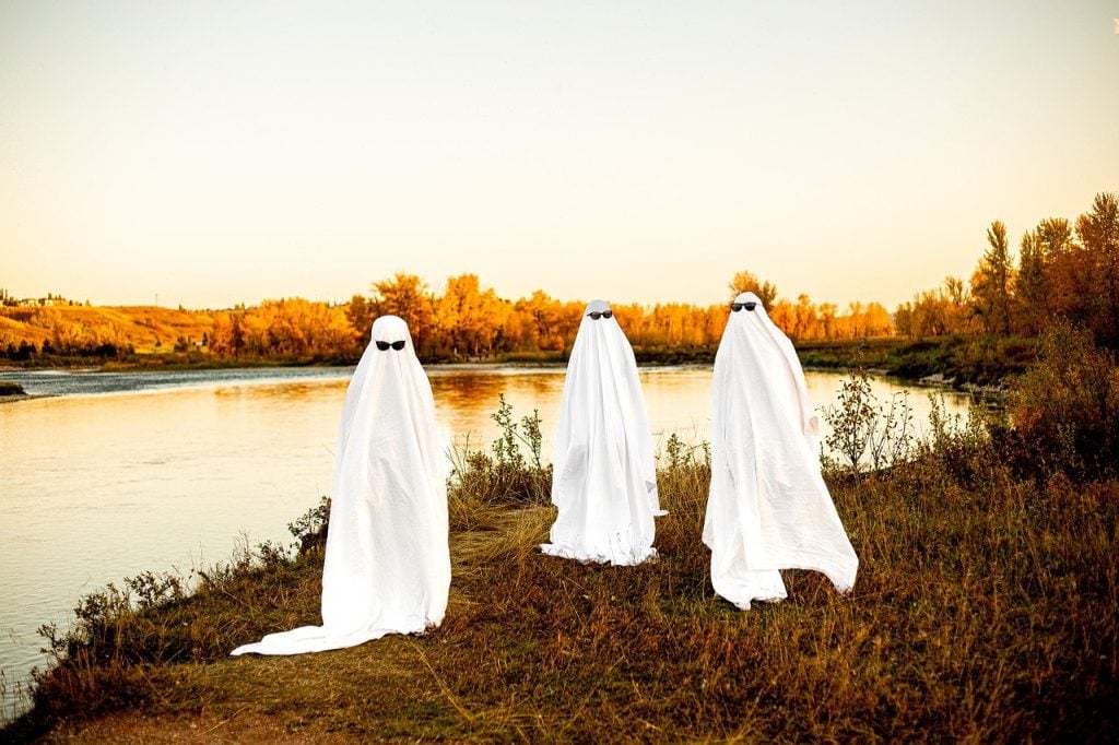 Three people in sheet ghost costumes wearing sunglasses standing next to a lake.